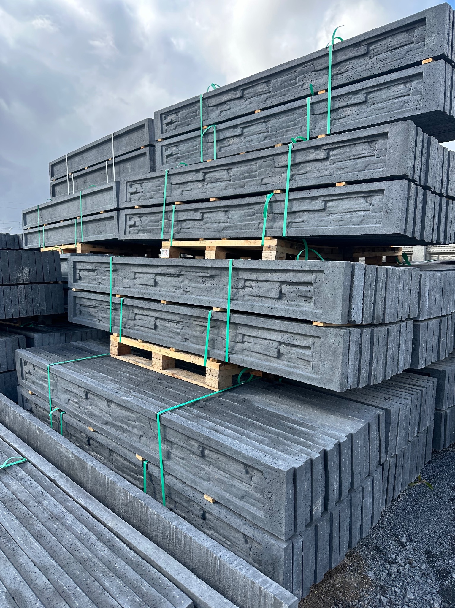 Szare, betonowe elementy ogrodzeniowe z fakturą kamienia, ułożone na paletach i zabezpieczone zielonymi taśmami. Widoczne detale faktury i struktury betonu.