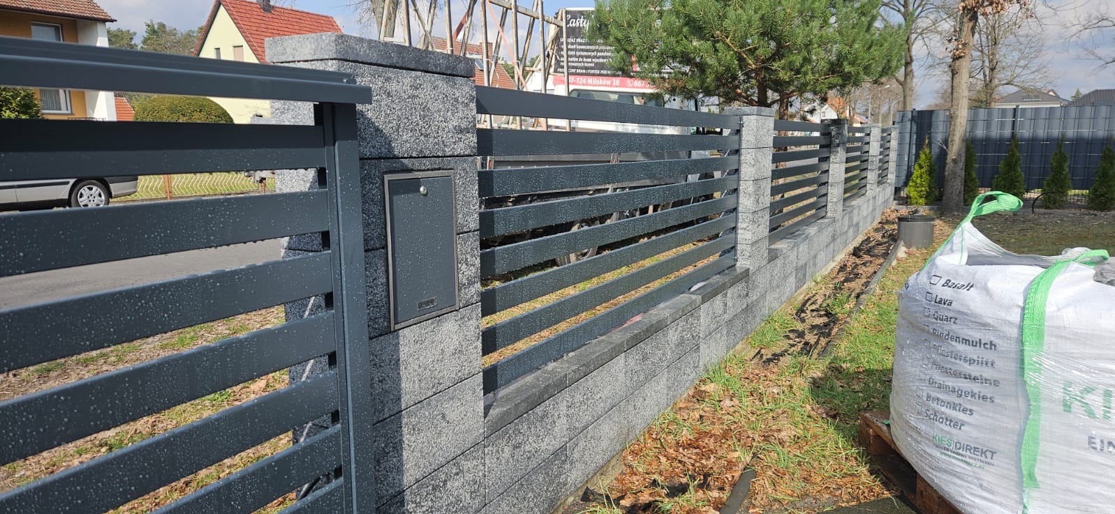 Nowoczesne ogrodzenie panelowe z szarego metalu i betonowych słupków, z zamontowaną skrzynką na listy. Widoczny worek z materiałami budowlanymi.