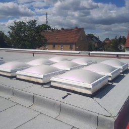 Arkadiusz Biegun - Szary dach pokryty papą z zamontowanymi świetlikami dachowymi. W tle budynki mieszkalne i błękitne niebo z chmurami.