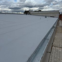 Arkadiusz Biegun - Szary, płaski dach z metalową rynną, widok z góry. W tle rusztowanie i pochmurne niebo. Minimalistyczny design dachu.