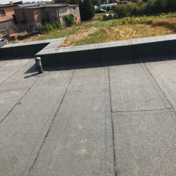 Arkadiusz Biegun - Szczegółowe ujęcie płaskiego dachu pokrytego papą termozgrzewalną z kominkiem wentylacyjnym. W tle zabudowa jednorodzinna i pole.