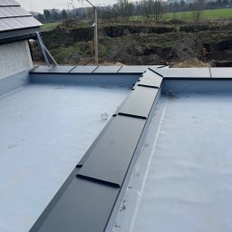 Arkadiusz Biegun - Szary dach płaski z czarnymi obróbkami blacharskimi, widok z góry na tle krajobrazu z budową i zielenią. Nowoczesne detale wykończenia dachu.