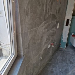 F H U Dariusz Kunicki - Szare, matowe płytki imitujące kamień na ścianie i podłodze w łazience w trakcie prac wykończeniowych. Widoczne przyłącza wodne i okno.