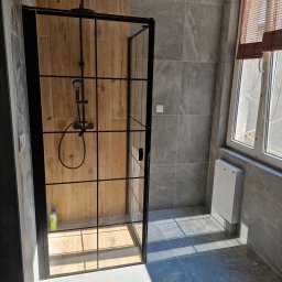 F H U Dariusz Kunicki - Nowoczesna kabina prysznicowa z czarnymi profilami i drewnopodobnymi płytkami wewnątrz, szare płytki na ścianach i podłodze, okno z drewnianymi żaluzjami.