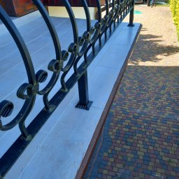 F H U Dariusz Kunicki - Kute, czarne balustrady balkonowe z ozdobnymi elementami w Nakle nad Notecią. Jasne płytki na balkonie i kolorowa kostka brukowa na podjeździe.