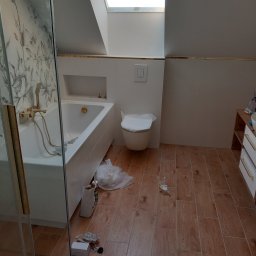 F H U Dariusz Kunicki - Wykończona łazienka na poddaszu z drewnopodobną podłogą, białą wanną z złotą armaturą, toaletą i oknem dachowym. Widoczne ślady po remoncie.
