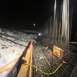 Ziolopol - Nocne ujęcie zbrojenia fundamentów pod budowę, z widocznym oświetleniem placu budowy i częściowo zasypanym śniegiem terenem. W tle koparka.