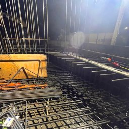 Ziolopol - Zbrojenie fundamentów z widoczną konstrukcją szalunkową i wystającymi prętami. W tle widoczny sprzęt budowlany i oświetlenie placu budowy.