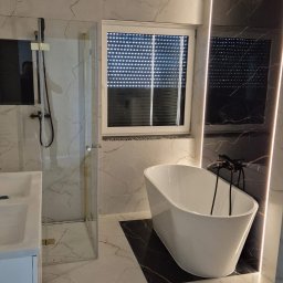 Marcin Buchowiecki - Nowoczesna łazienka z marmurowymi płytkami, wolnostojącą wanną i kabiną prysznicową ze złotymi akcentami. Oświetlenie LED podkreśla elegancki design.