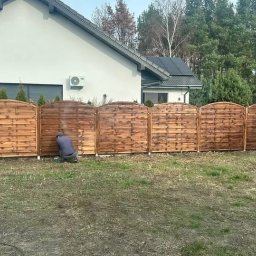 Marcin Buchowiecki - Malowanie drewnianego ogrodzenia panelowego na zewnątrz domu. Widoczny fragment pomalowany i osoba w trakcie pracy. Trawa i drzewa w tle.