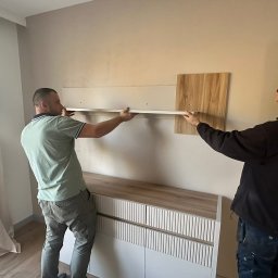 Marcin Buchowiecki - Dwóch monterów instaluje półkę na ścianie nad komodą w jasnym wnętrzu. Widoczne narzędzia i elementy montażowe. Praca w toku, etap montażu.