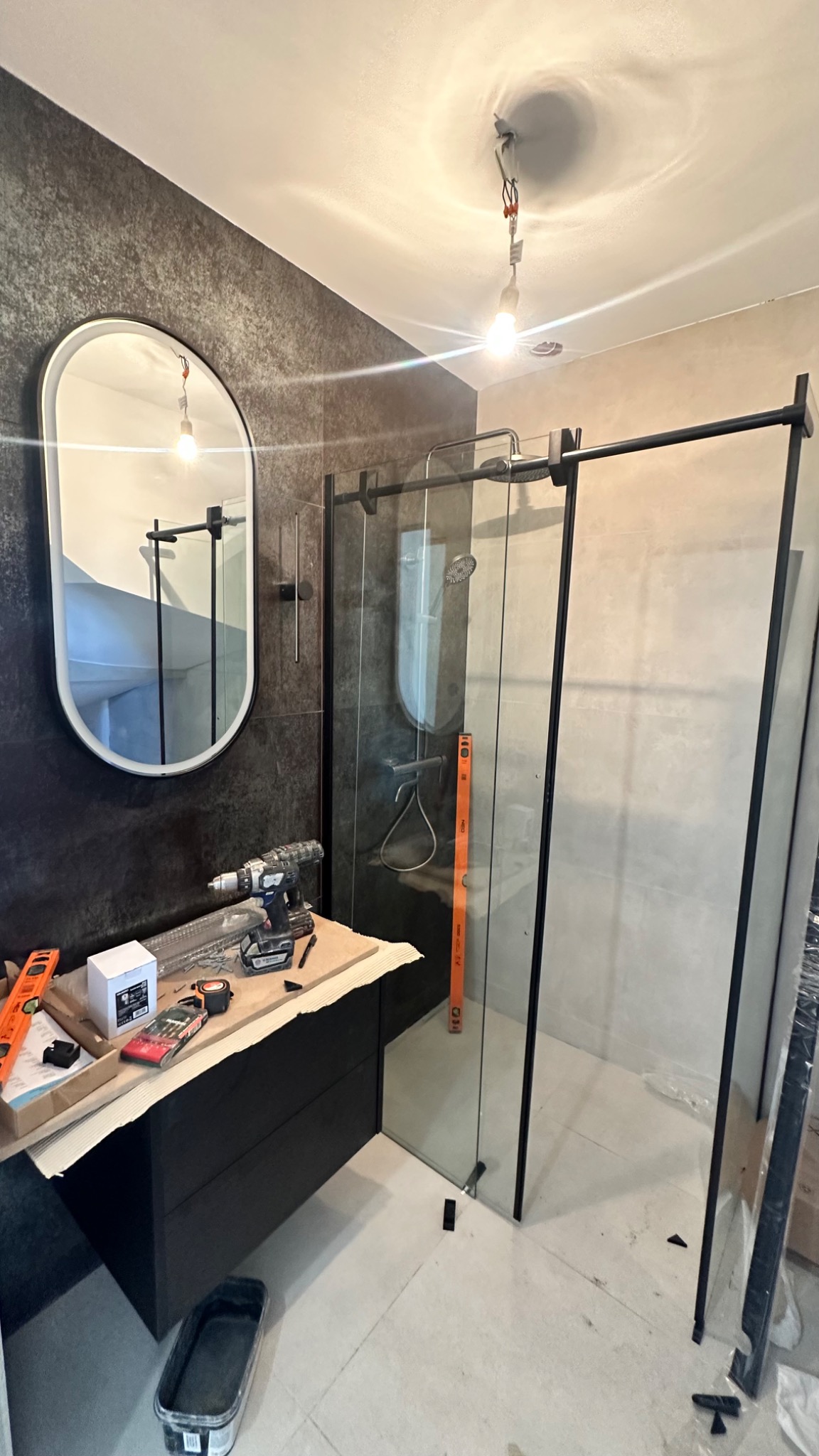 Nowoczesna łazienka w trakcie wykańczania: czarna szafka z umywalką, owalne lustro i kabina prysznicowa z czarnymi ramami. Widoczne narzędzia i poziomica.