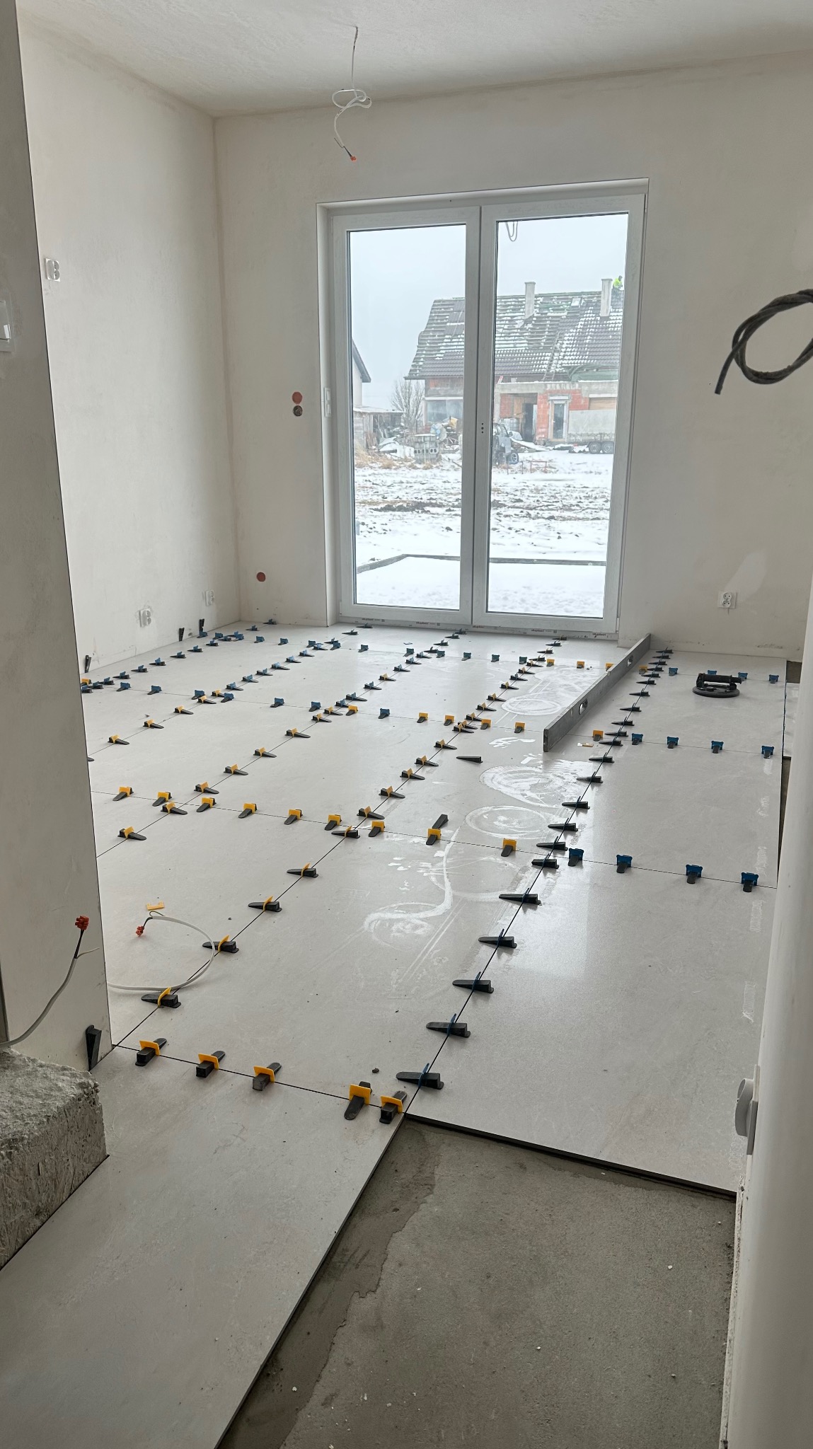 Jasne płytki podłogowe w trakcie układania, z systemem poziomowania. Widoczne narzędzia glazurnicze i okno z widokiem na zimowy krajobraz. Wnętrze w stanie surowym.