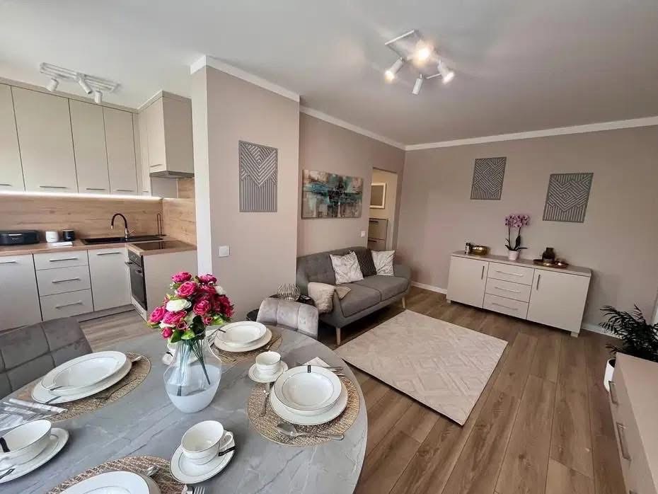 Jasne, beżowe wnętrze po remoncie: kuchnia z drewnianym blatem, szary narożnik, stół z zastawą i drewniana podłoga. Dekoracyjne panele ścienne.