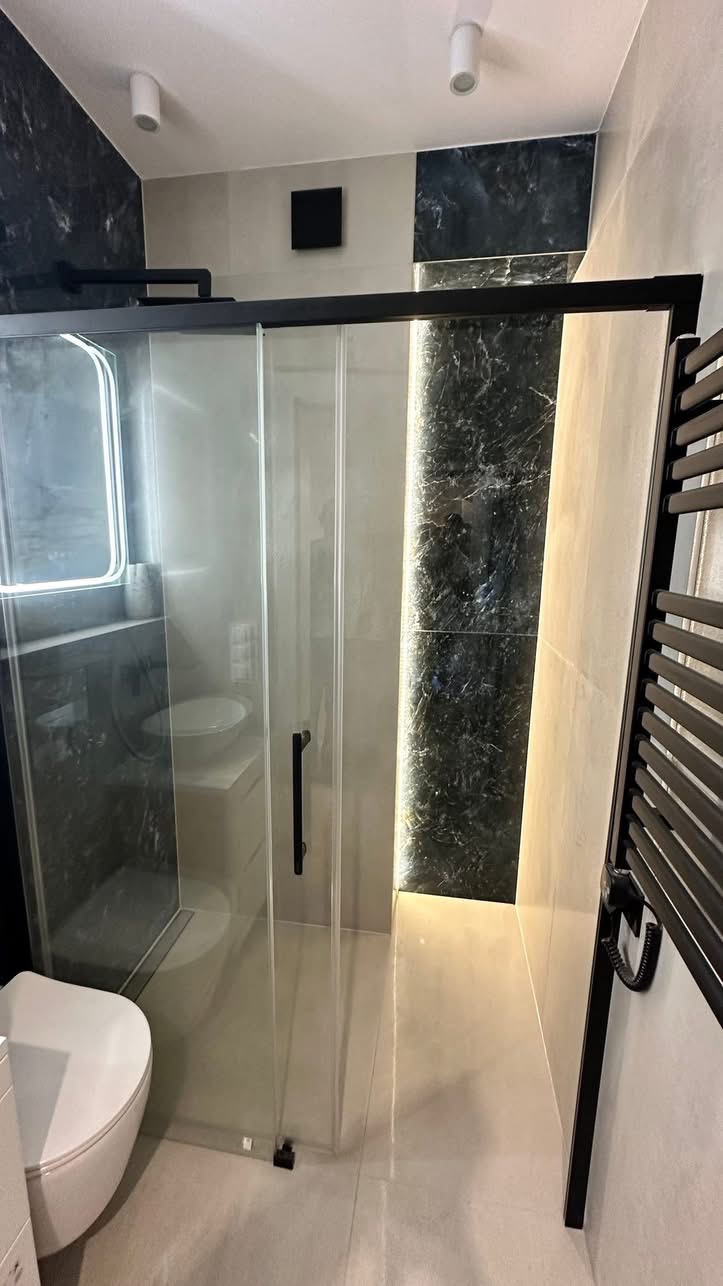 Nowoczesna łazienka z kabiną prysznicową typu walk-in z czarnymi detalami, marmurowymi akcentami i grzejnikiem drabinkowym. Minimalistyczny design, jasne płytki podłogowe.