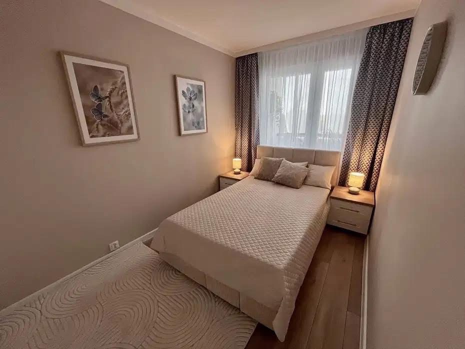 Przytulna sypialnia w beżach: podwójne łóżko, drewniana podłoga, nocne lampki i dekoracyjne obrazy na ścianie. Subtelne zasłony w oknie.