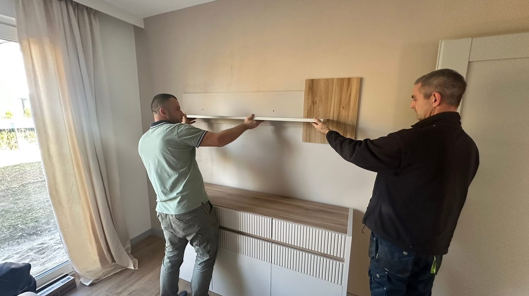 Dwóch monterów instaluje półkę na ścianie nad komodą w jasnym wnętrzu. Widoczne narzędzia i elementy montażowe. Praca w toku, etap montażu.