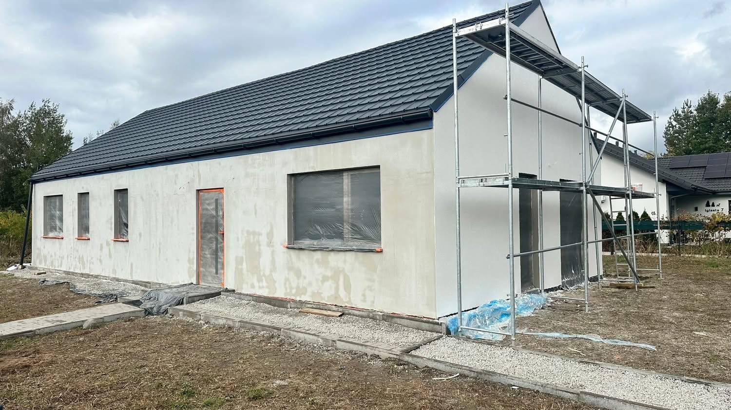 Dom jednorodzinny w trakcie szpachlowania elewacji, okna i drzwi zabezpieczone folią, widoczne rusztowanie. Prace wykończeniowe na zewnątrz budynku.