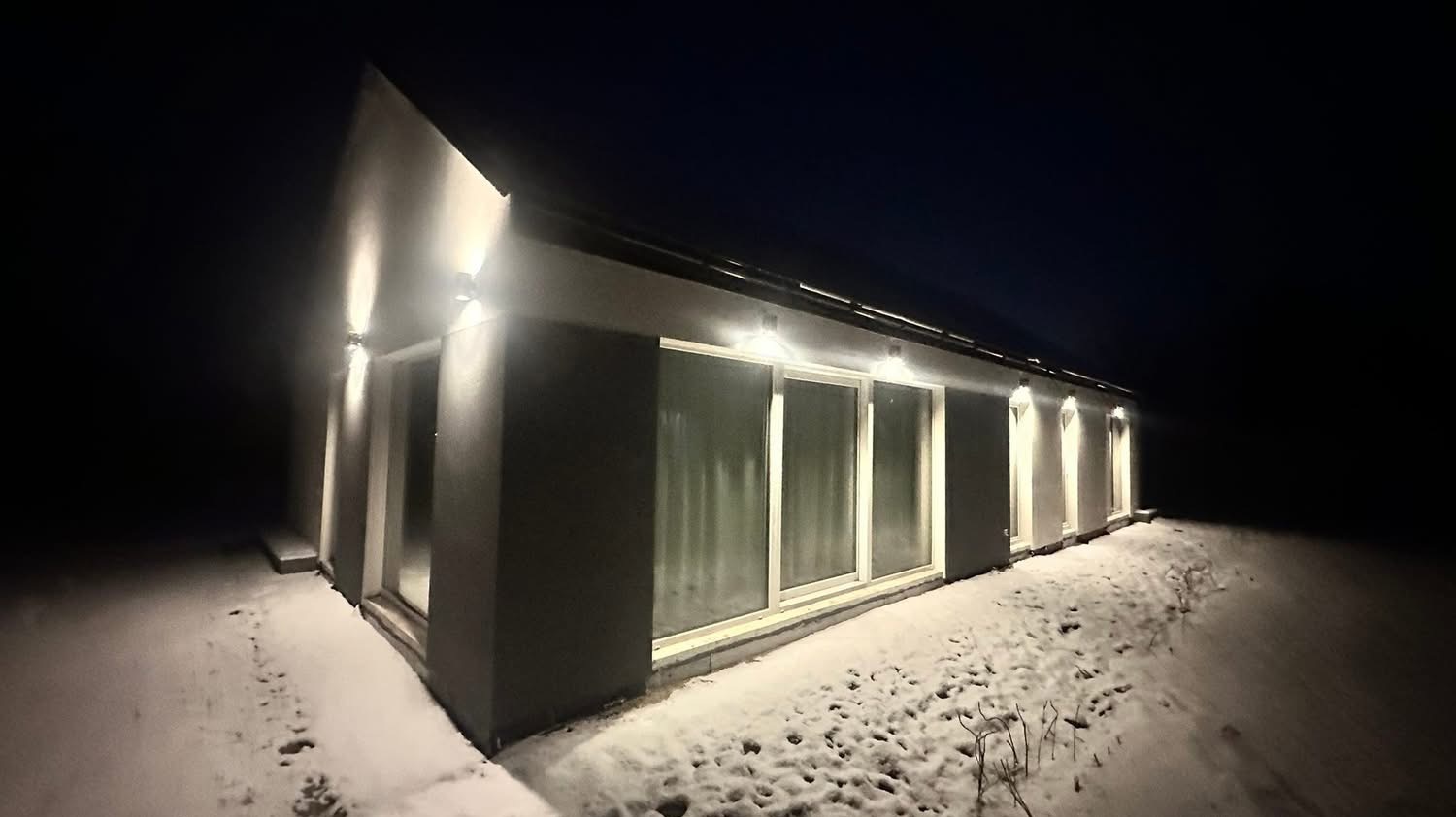 Nowoczesny dom jednorodzinny nocą, oświetlony zewnętrznymi lampami ściennymi, z widocznym śniegiem na ziemi. Minimalistyczna architektura z dużymi oknami.