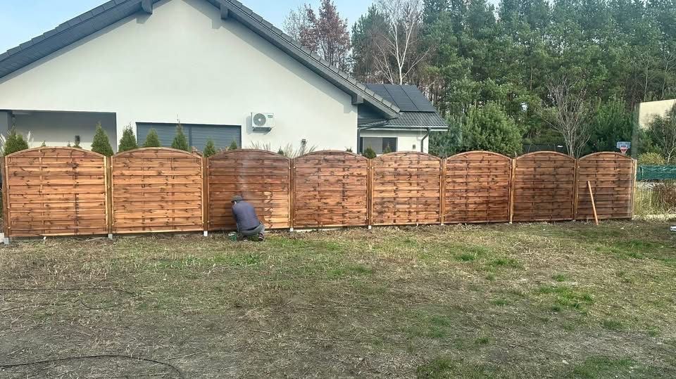 Malowanie drewnianego ogrodzenia panelowego na tle domu z szarą dachówką. Widoczny pracownik w trakcie pracy, używający pistoletu natryskowego. Świeżo pomalowane panele.