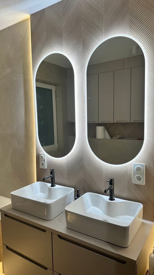 Nowoczesna łazienka z dwoma białymi umywalkami nablatowymi, czarnymi bateriami i podświetlanymi lustrami owalnymi na ścianie z wzorem jodełki.