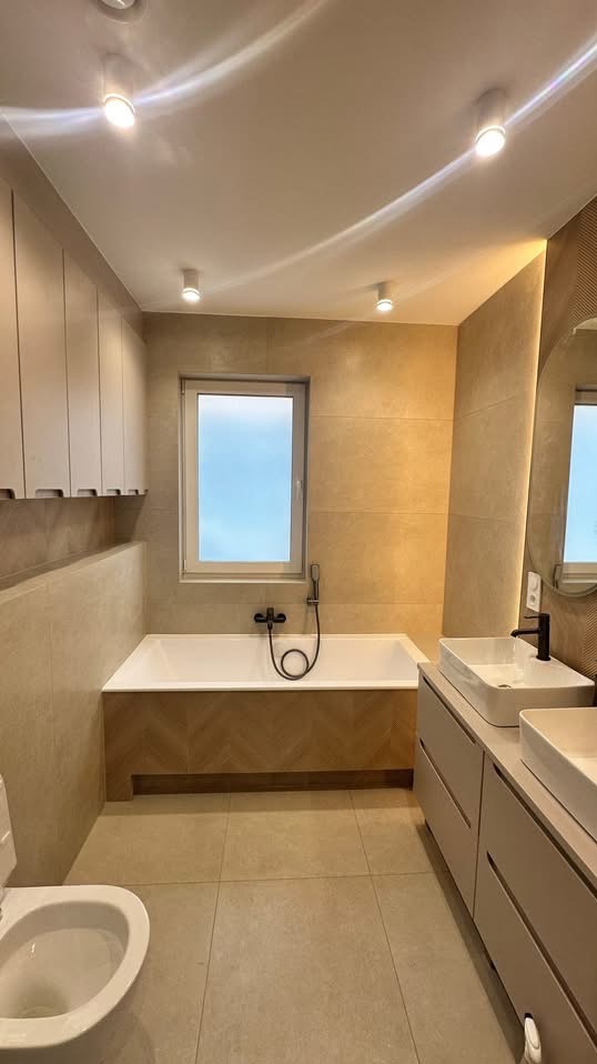 Jasna łazienka z beżowymi płytkami, białą wanną obudowaną drewnem w jodełkę, umywalkami nablatowymi i szafkami. Minimalistyczny design, oświetlenie punktowe.