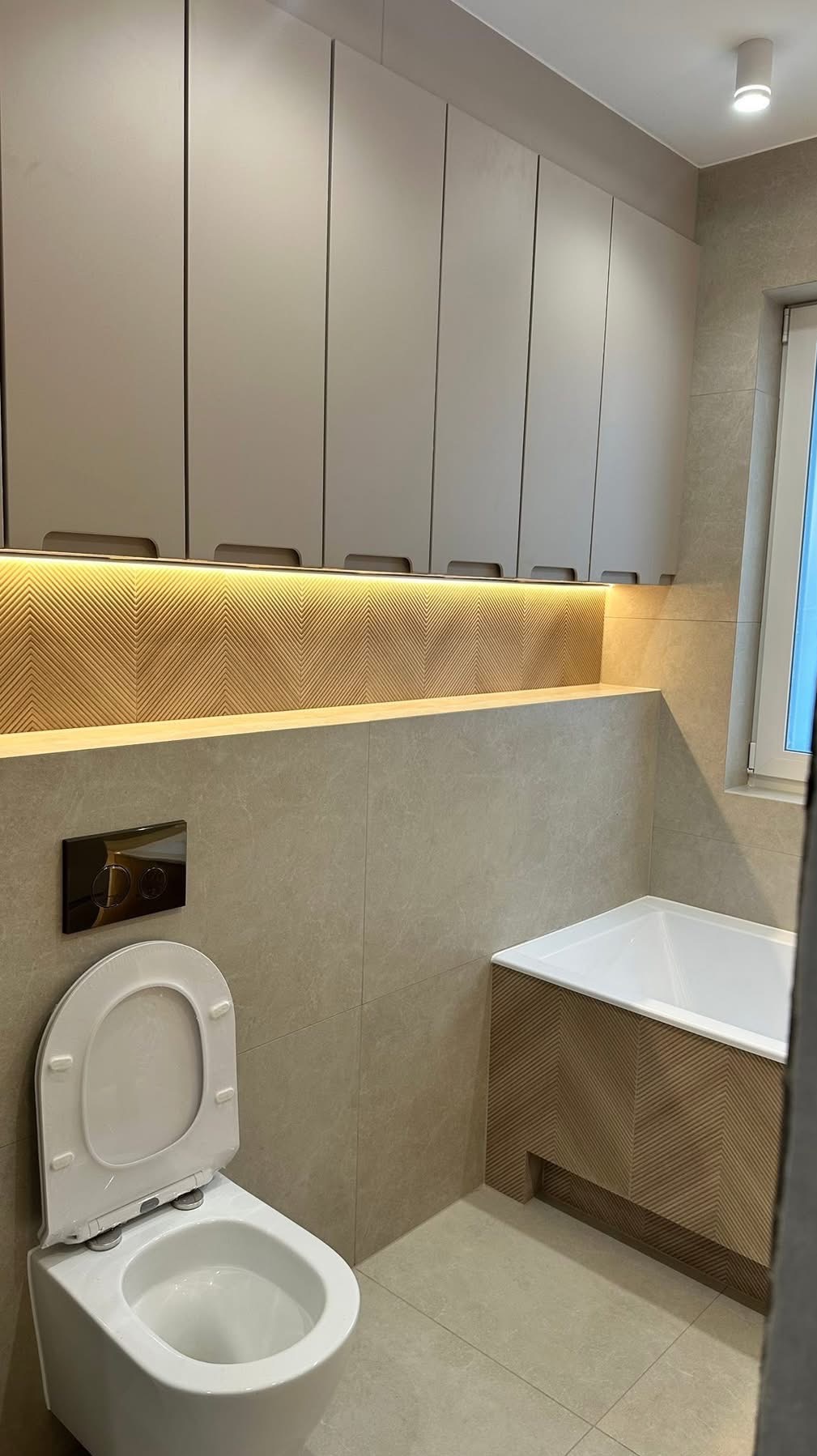 Nowoczesna łazienka w beżach: podwieszana toaleta, wanna z obudową w jodełkę, szafki wiszące z oświetleniem LED. Minimalistyczny design, płytki imitujące kamień.