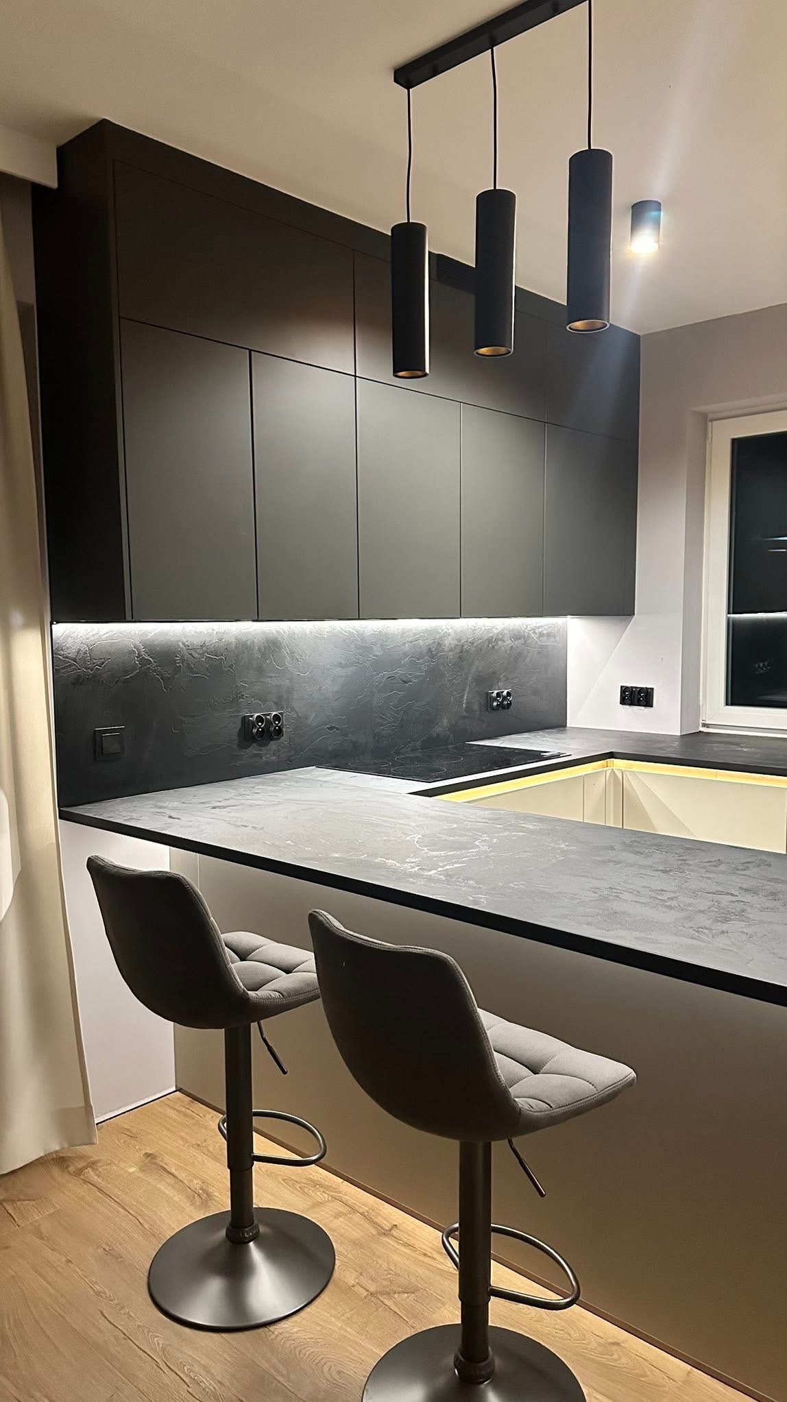 Nowoczesna kuchnia w odcieniach szarości z podświetleniem LED, wyspą i hokerami. Minimalistyczne lampy wiszące. Wykończenie wnętrz z dbałością o detale.