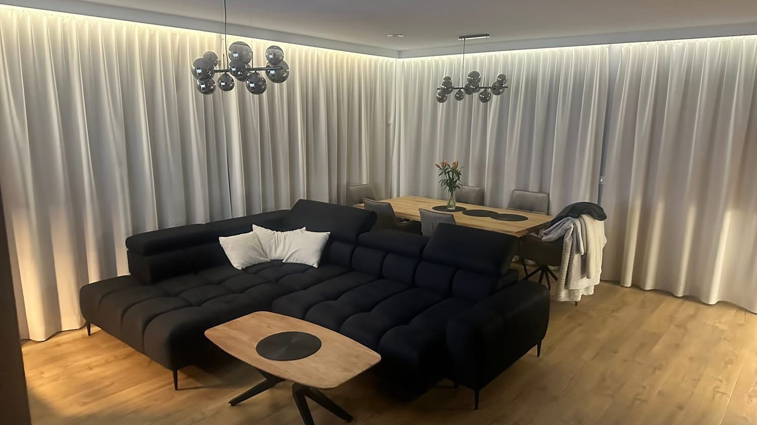 Nowoczesny salon z czarną sofą modułową, drewnianym stolikiem kawowym i stołem jadalnym w tle, oświetlony lampami sufitowymi i listwami LED.