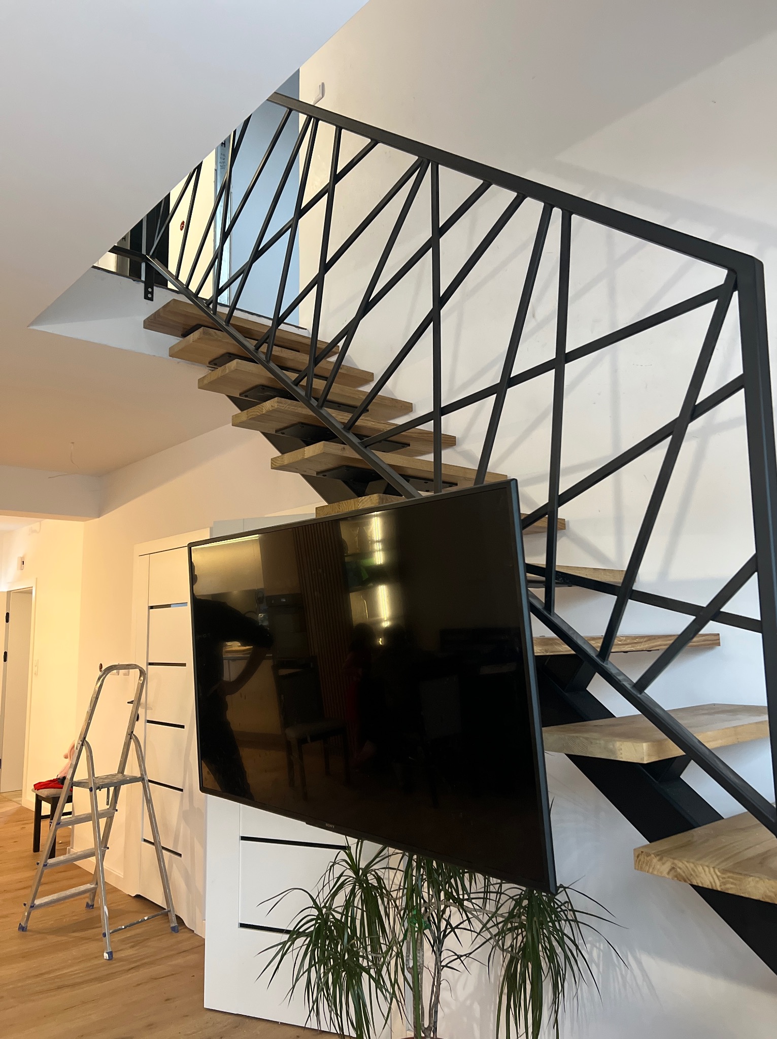 Nowoczesne schody z drewnianymi stopniami i czarną, geometryczną balustradą. W tle drabina i szafka RTV z telewizorem. Minimalistyczny design wnętrza.
