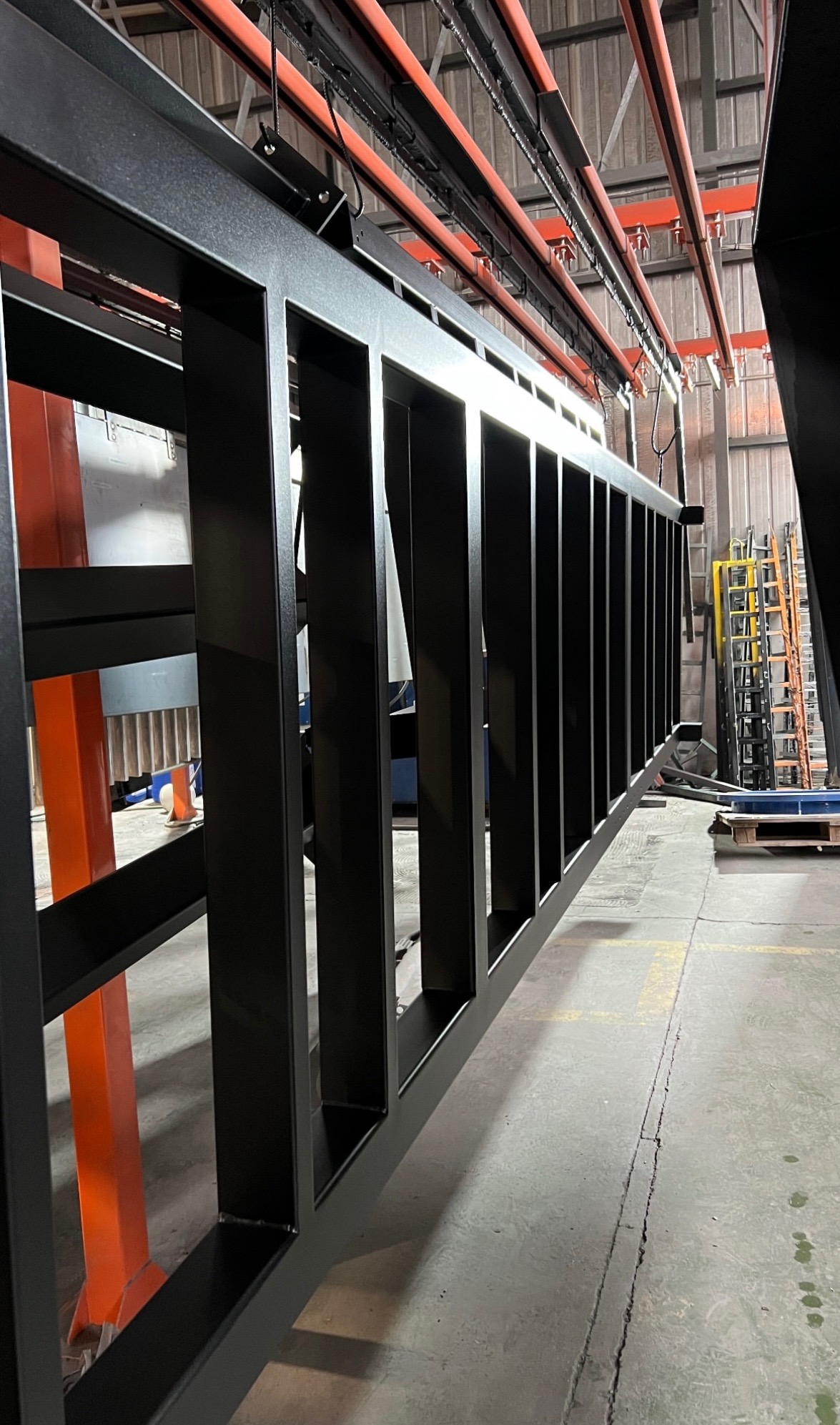 Czarna, metalowa konstrukcja balustrady w hali produkcyjnej, zawieszona na pomarańczowych elementach transportowych. Widoczne spawane łączenia i surowy beton na podłodze.