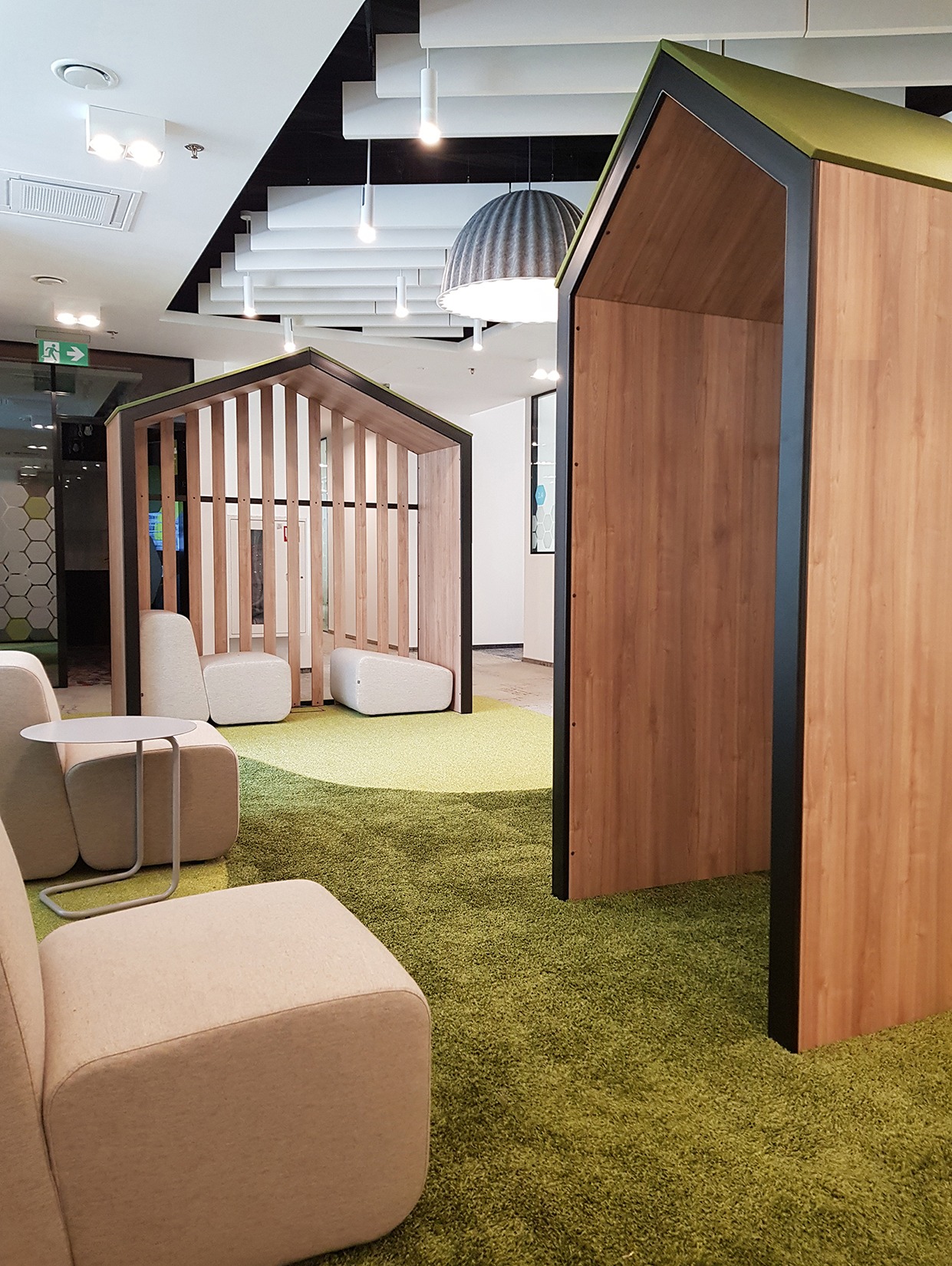 Nowoczesna aranżacja biura z zieloną wykładziną i drewnianymi kabinami w kształcie domków, tworzącymi strefę relaksu. Widoczne minimalistyczne meble i oświetlenie.