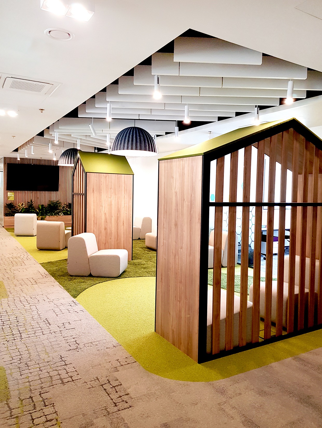 Nowoczesna aranżacja biura z drewnianymi kabinami w kształcie domków, zieloną wykładziną i designerskimi lampami. Przestrzeń relaksu i pracy w otoczeniu roślin.