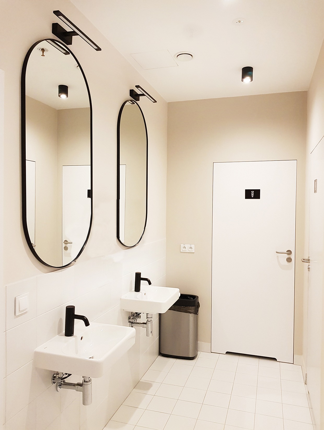 Nowoczesna łazienka publiczna z dwoma umywalkami, owalnymi lustrami w czarnych ramach i minimalistycznym oświetleniem. Białe płytki i drzwi, metalowy kosz na śmieci.