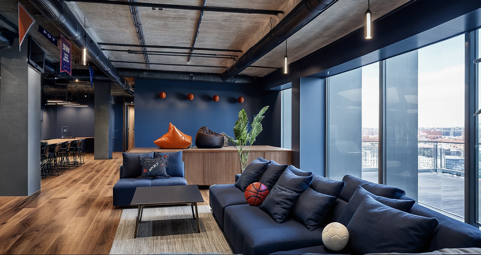 Nowoczesne biuro z strefą relaksu: granatowe ściany, drewniana podłoga, sofy z poduszkami, pufy, piłki do koszykówki i piłka nożna. Duże okna z widokiem na miasto.