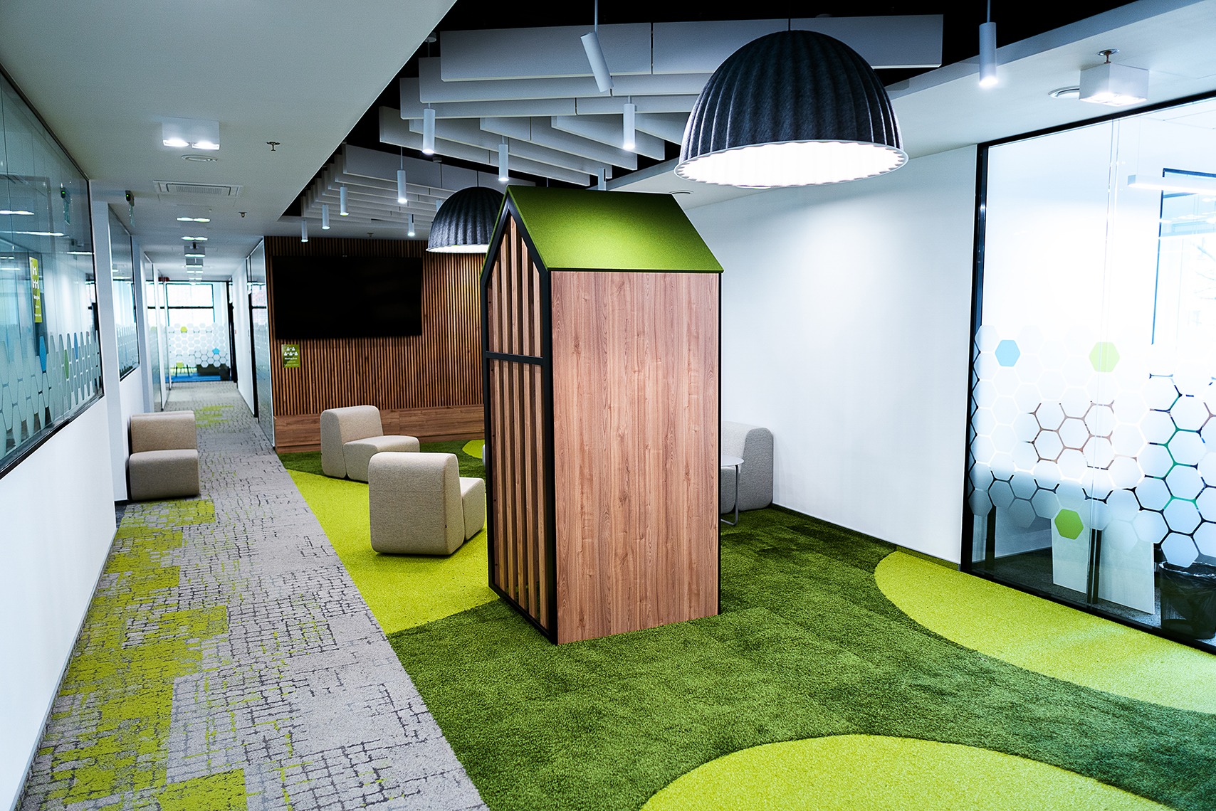Nowoczesna aranżacja biura: strefa relaksu z drewnianą budką i zielonym dywanem, jasne krzesła, przeszklone ściany z dekoracyjnymi wzorami, designerskie lampy.