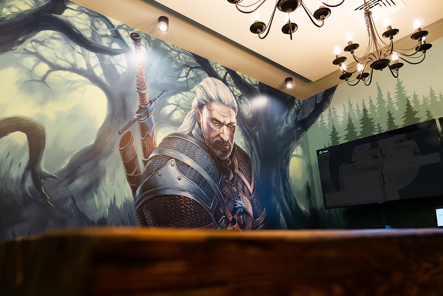 Mural z wizerunkiem Geralta z Rivii na ścianie biura, oświetlony lampami sufitowymi, z żyrandolem i telewizorem w tle. Widok zza drewnianego blatu.