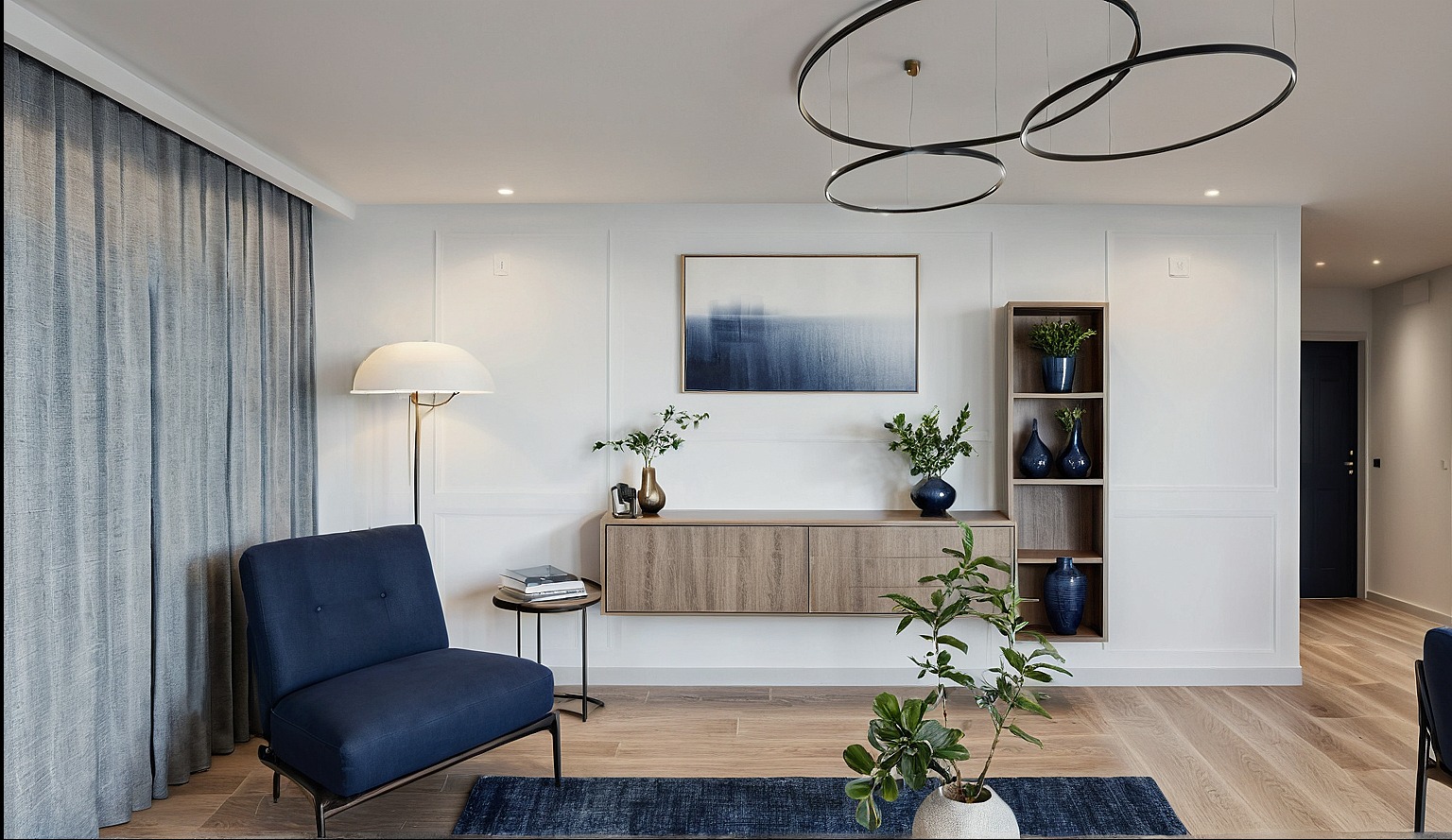Nowoczesny salon z niebieskim akcentem: fotel, dywan i dekoracje. Jasne drewno, białe ściany z panelami, abstrakcyjny obraz i designerska lampa pierścieniowa.