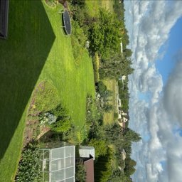 Monika Hnat - Zadbany trawnik z trampoliną i szklarnią w słoneczny dzień. Widok z góry na zielony ogród z kwitnącymi rabatami i drzewami. Błękitne niebo z białymi chmurami.