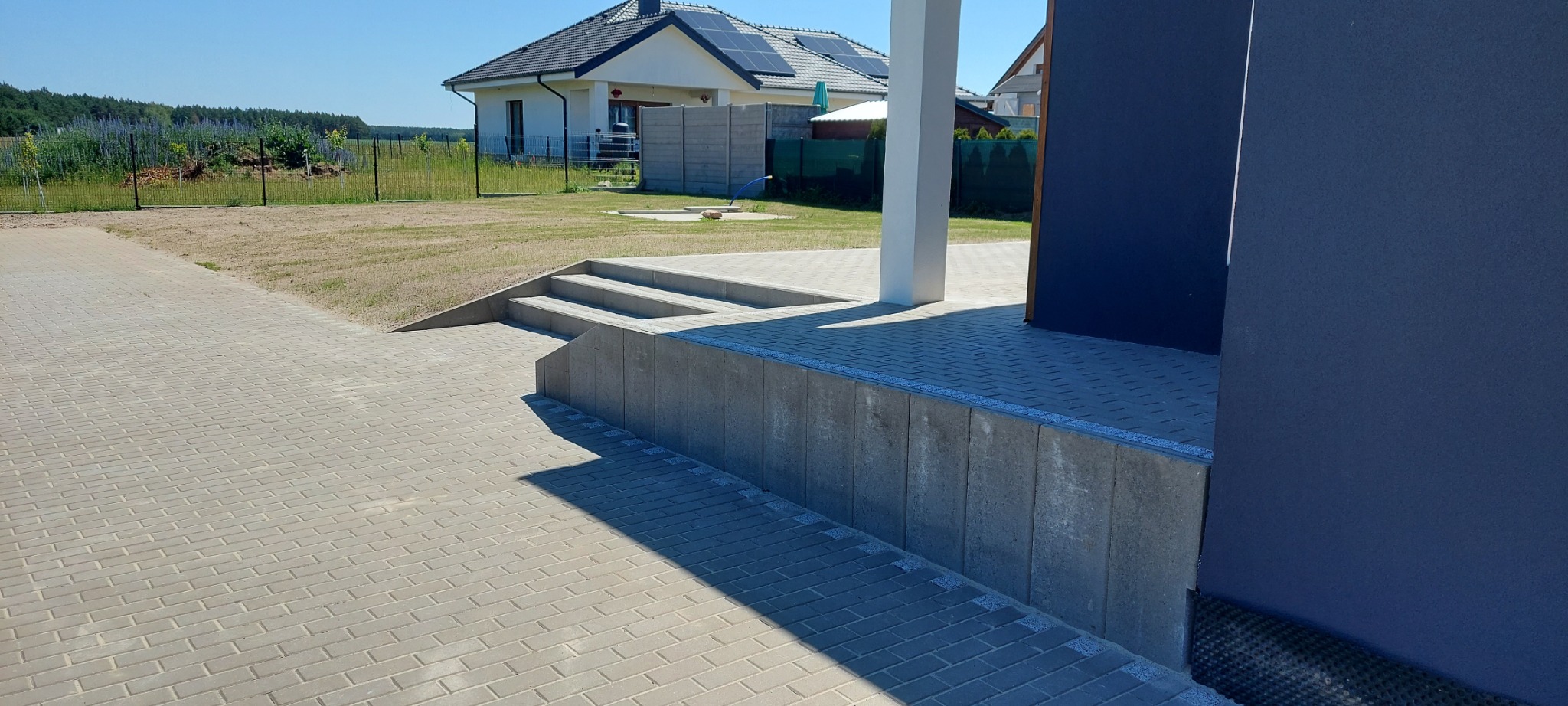Jasnoszara kostka brukowa ułożona przed nowoczesnym domem z panelami słonecznymi. Widoczne schody wejściowe i fragment elewacji w kolorze granatowym.