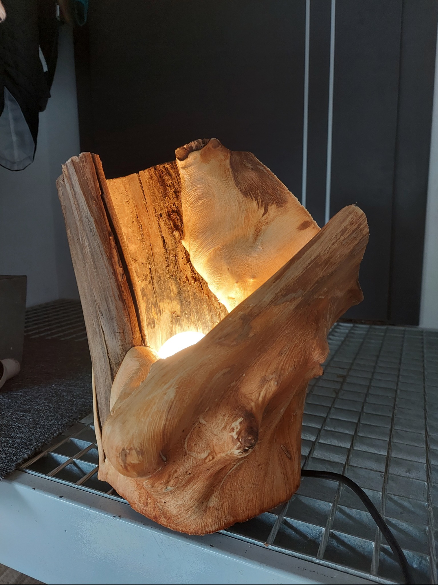 Unikatowa lampa z drewna, ręcznie wykonana, z widocznym kablem zasilającym. Ciepłe światło wydobywa naturalną strukturę drewna. Stoi na metalowej kratce.