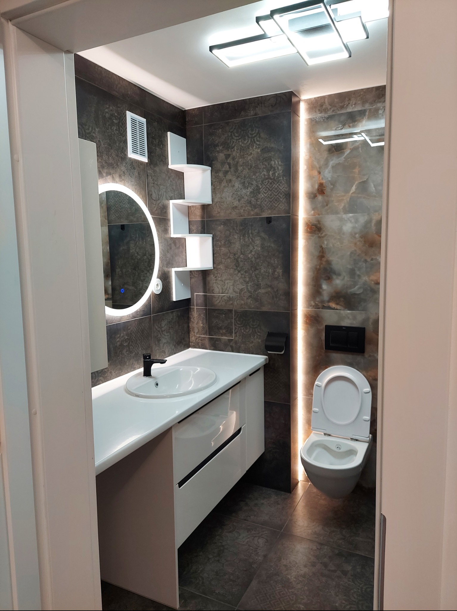Nowoczesna łazienka z brązowymi płytkami, białym blatem z umywalką, okrągłym lustrem z podświetleniem LED i podwieszaną toaletą z czarnym przyciskiem.