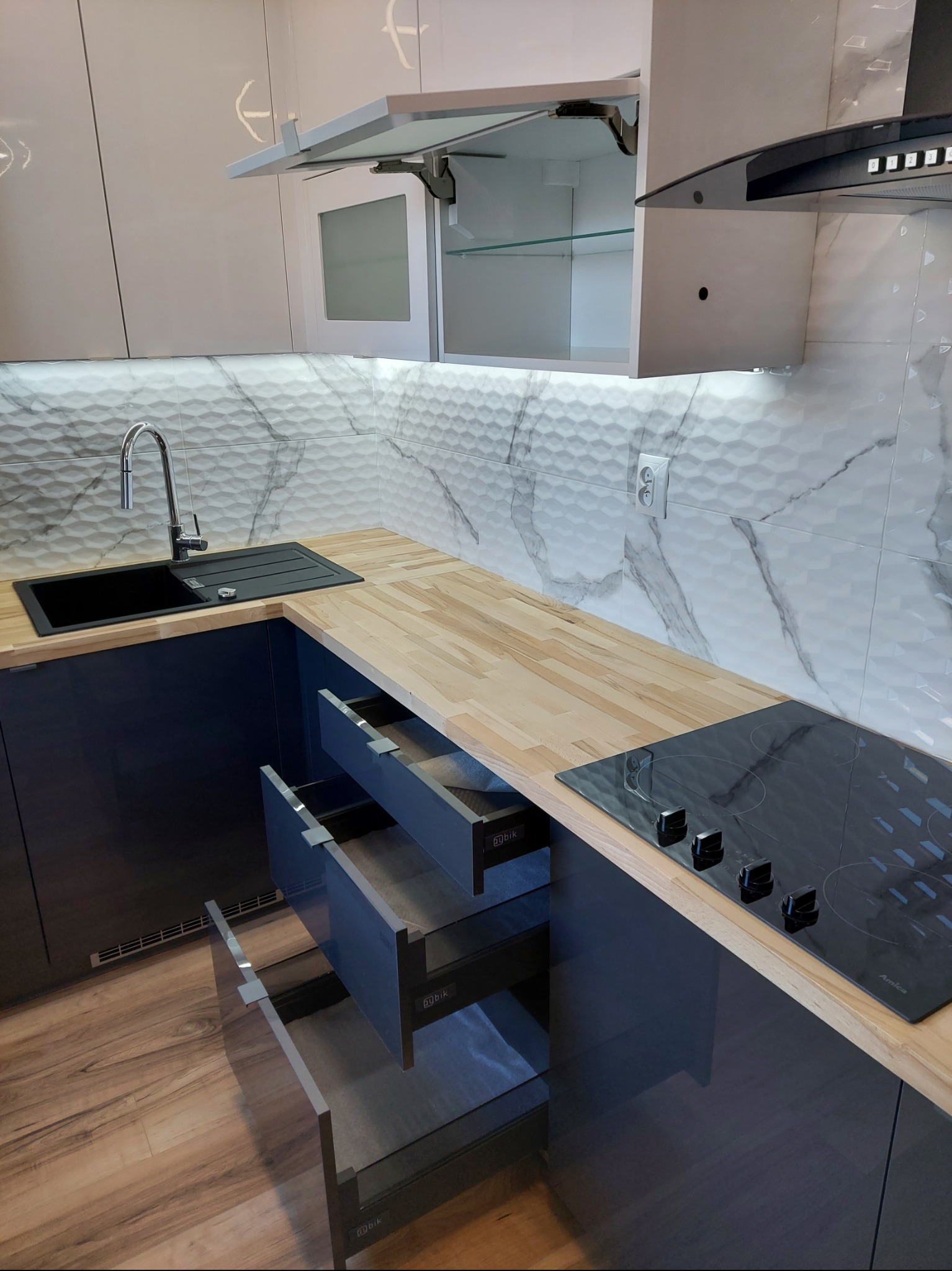 Nowoczesna kuchnia z drewnianym blatem, czarnym zlewem i płytą indukcyjną. Otwarta szafka górna i wysunięte szuflady ukazują detale wykończenia. Marmurowe płytki na ścianie.