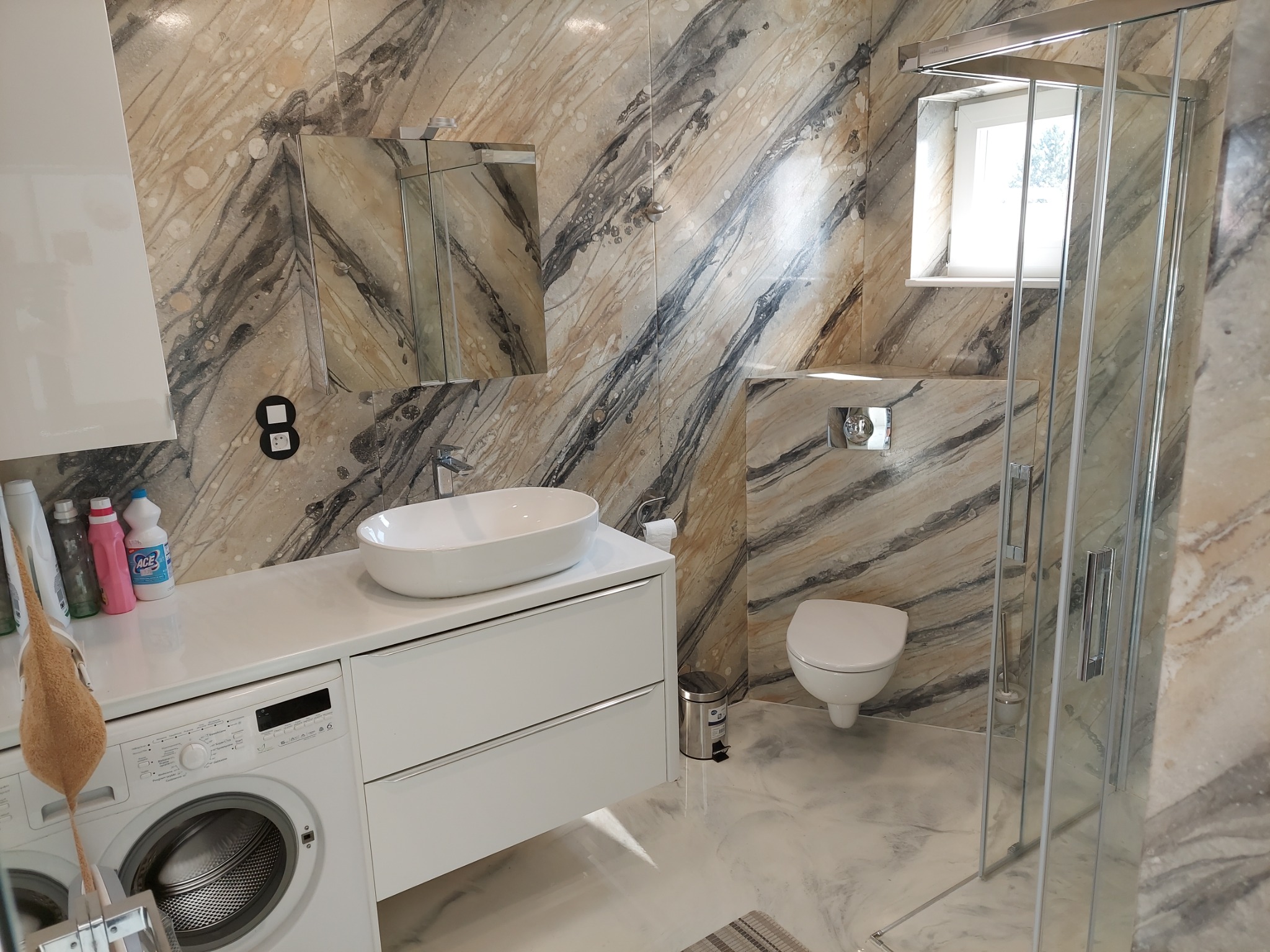 Wykończona łazienka z marmurowymi płytkami, białą umywalką nablatową, pralką pod blatem i kabiną prysznicową. Nowoczesny design z akcentami srebra.