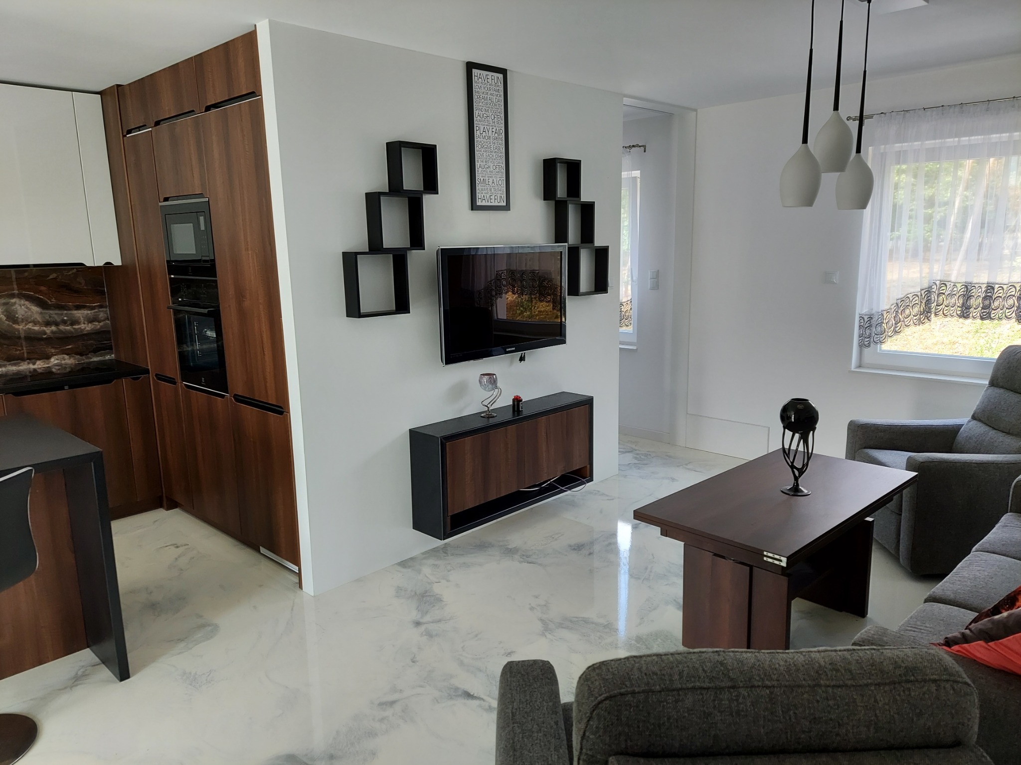 Jasne wnętrze z posadzką żywiczną imitującą marmur. Na ścianie telewizor otoczony czarnymi półkami. Widoczne meble kuchenne i wypoczynkowe w odcieniach brązu i szarości.
