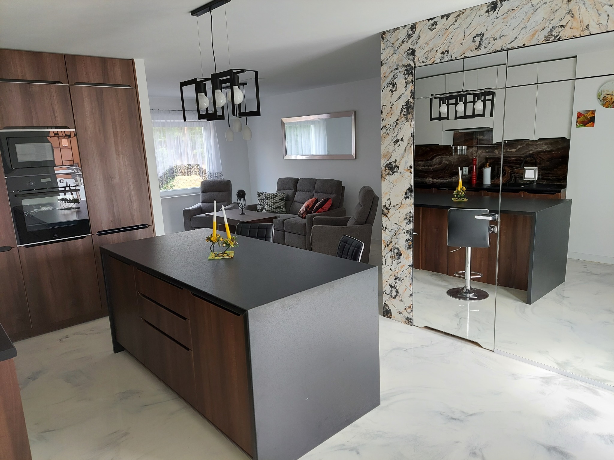 Nowoczesne wnętrze: wyspa kuchenna z ciemnym blatem, drewniane szafki, posadzka żywiczna imitująca marmur. W tle salon z szarą sofą i lustrem.
