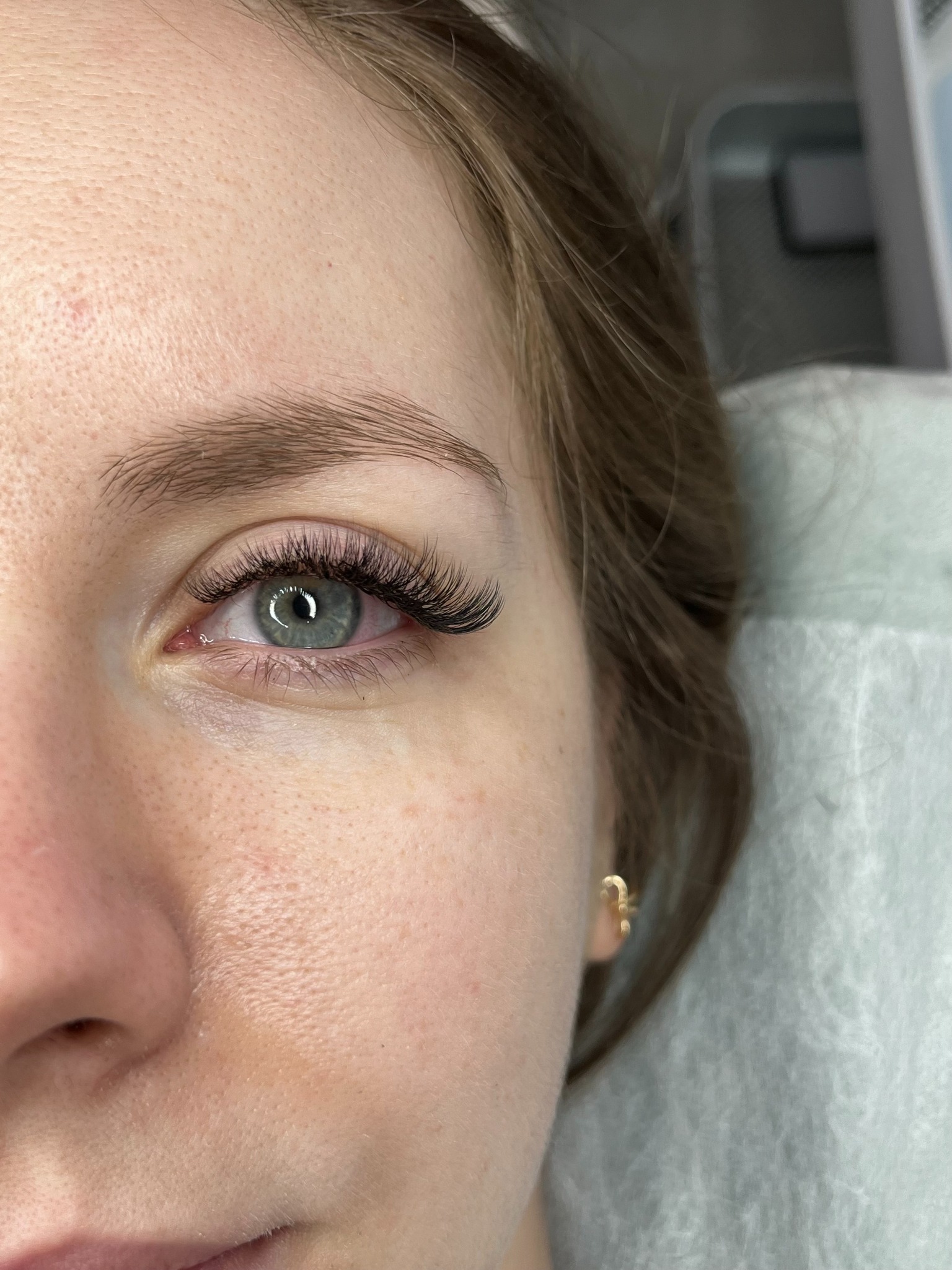 Zbliżenie na oko kobiety z profesjonalnie przedłużonymi rzęsami, widoczna złota kolczyk w uchu. Delikatny makijaż podkreśla naturalne piękno.