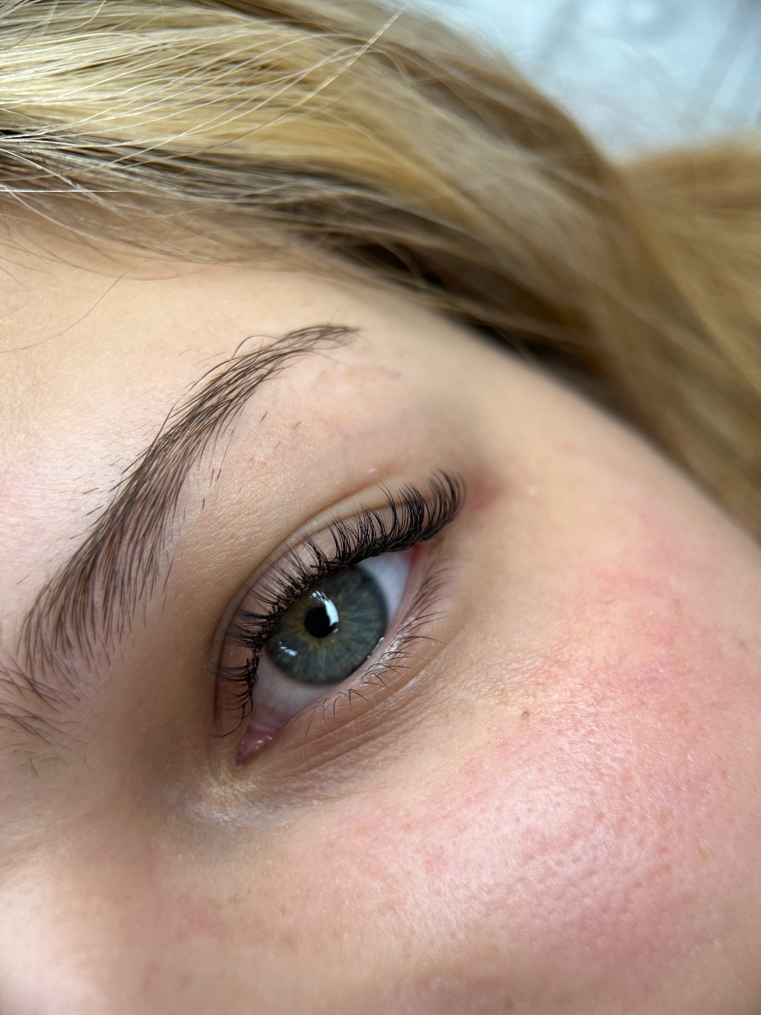 Zbliżenie na oko z przedłużonymi rzęsami, widoczne pojedyncze włoski, naturalny wygląd, brwi podkreślone, jasna karnacja, blond włosy w tle.