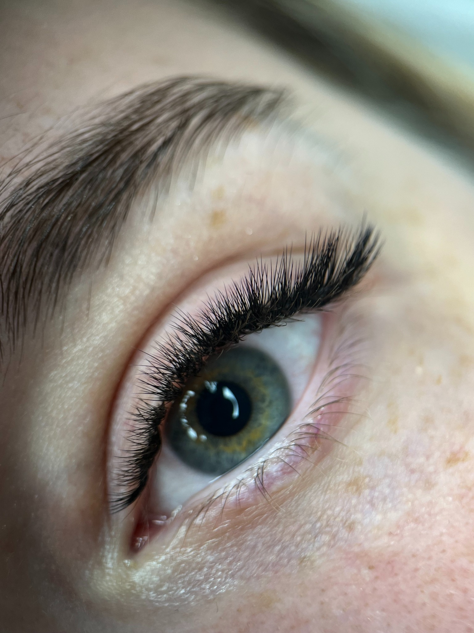 Zbliżenie na oko z wyraźnie przedłużonymi, czarnymi rzęsami. Widoczne piegi na skórze i zielono-szary kolor tęczówki. Makro ujęcie podkreśla precyzję zabiegu.