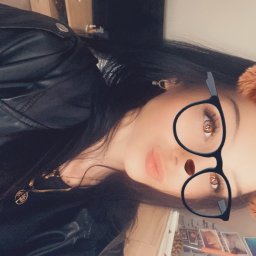 Dominika Behrendt - Portret kobiety w czarnej kurtce, z filtrem okularów i nosa. W tle widoczny regał i ściana z naklejkami. Nietypowy kadr, obrócony o 90 stopni.